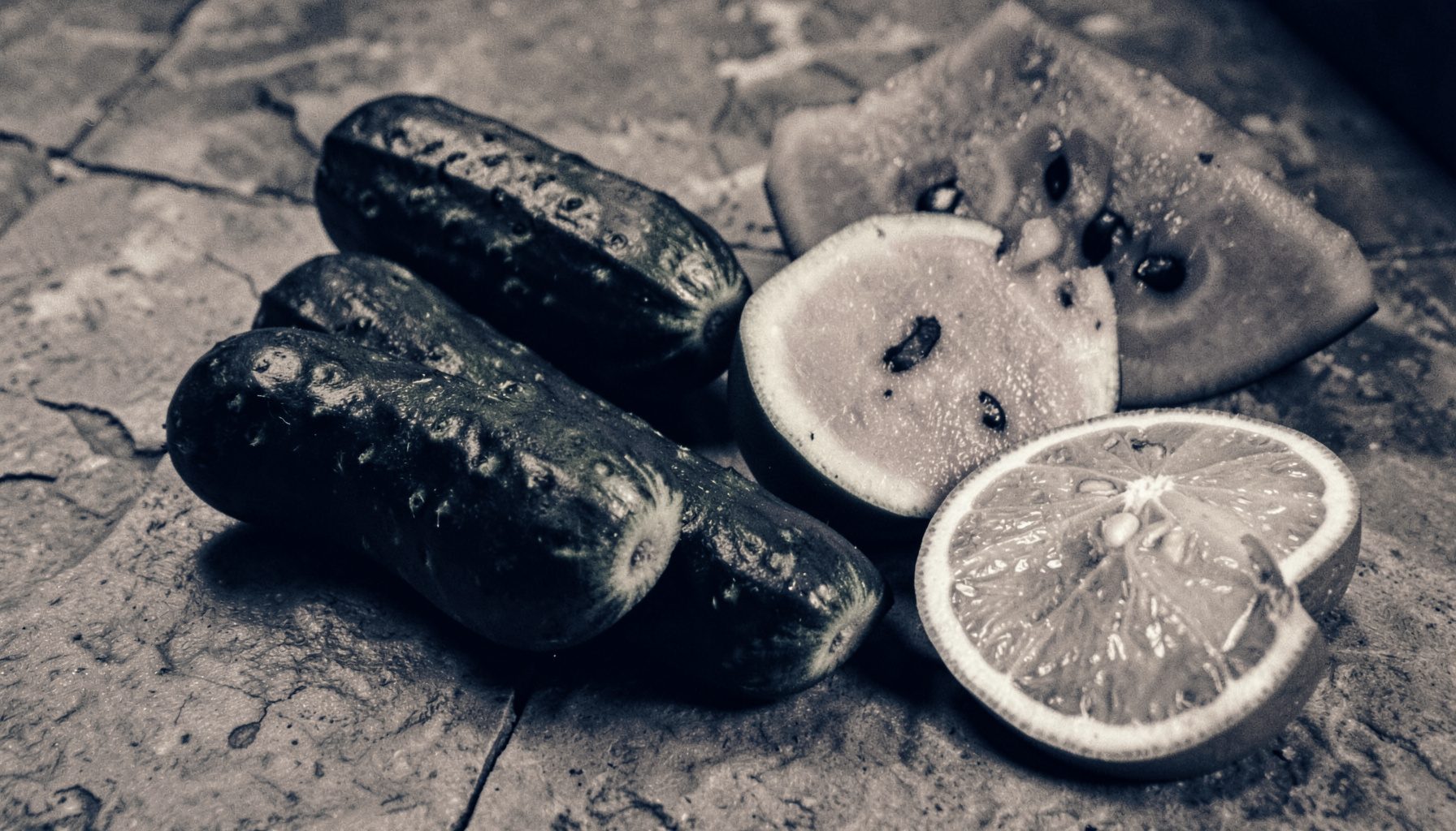 A duotone photograph of crisp cucumbers, watermelon slices, and citrus fruits on a cool stone surface, rich amber and charcoal tones