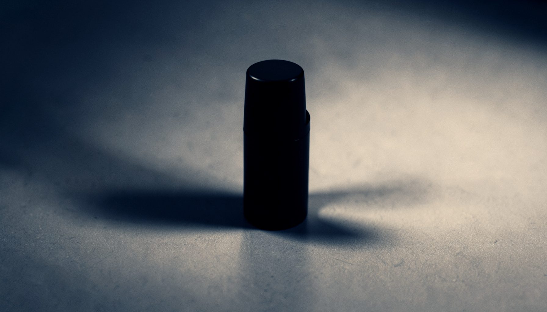 A duotone still life of a black lipstick tube resting on a matte surface, dramatic lighting with deep charcoal and warm ivory tones.