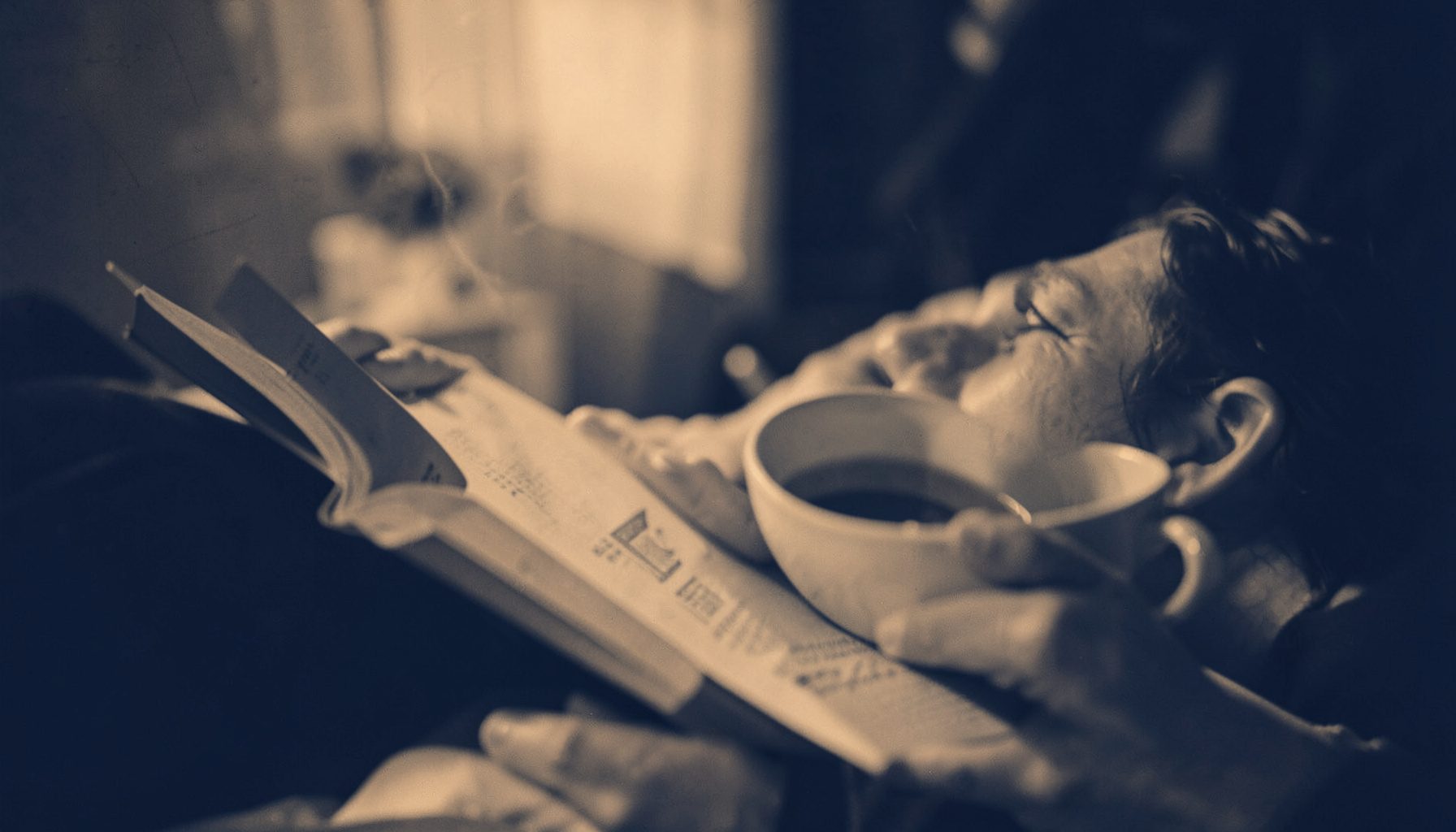 A warm duotone photograph of a person relaxing with a book and a cup of tea, soft amber and charcoal tones