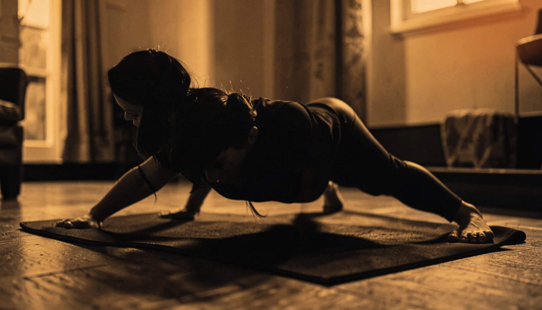 A duotone photograph of a person performing a bodyweight exercise on a mat at home, bathed in warm amber and deep charcoal