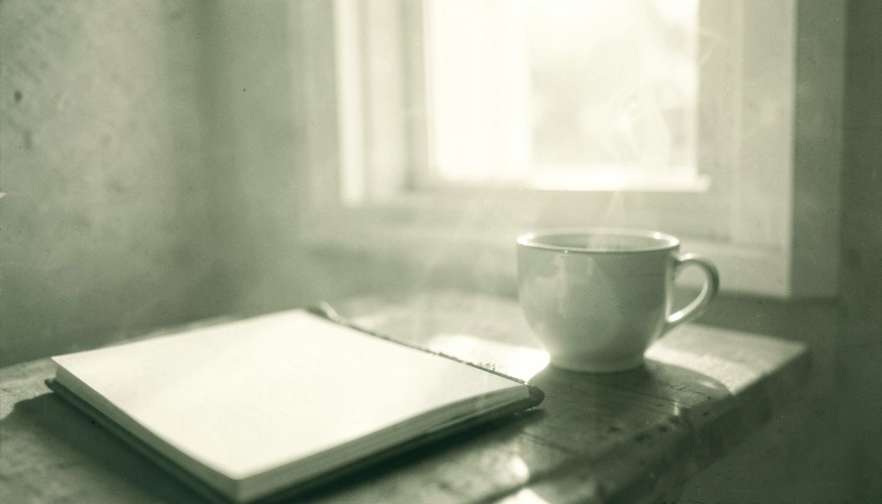 A duotone photograph of a peaceful morning scene with a journal, a cup of tea, and soft sunlight streaming through a window