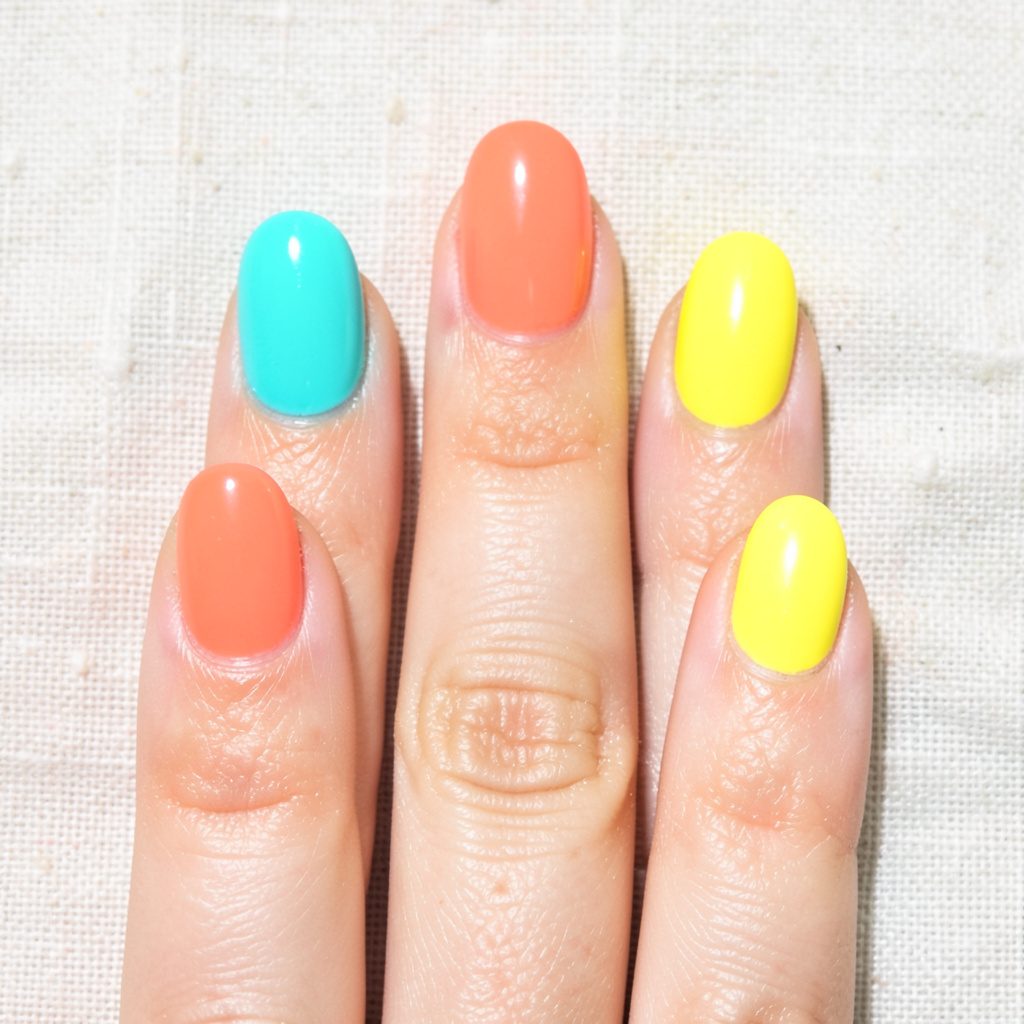 Five freshly painted fingernails in vivid coral, turquoise, and sunny yellow shades against a light linen background