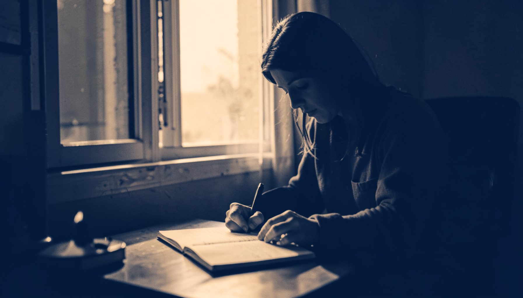 A duotone photograph of a person journaling at a desk by a window with soft morning light, warm amber and deep navy tones